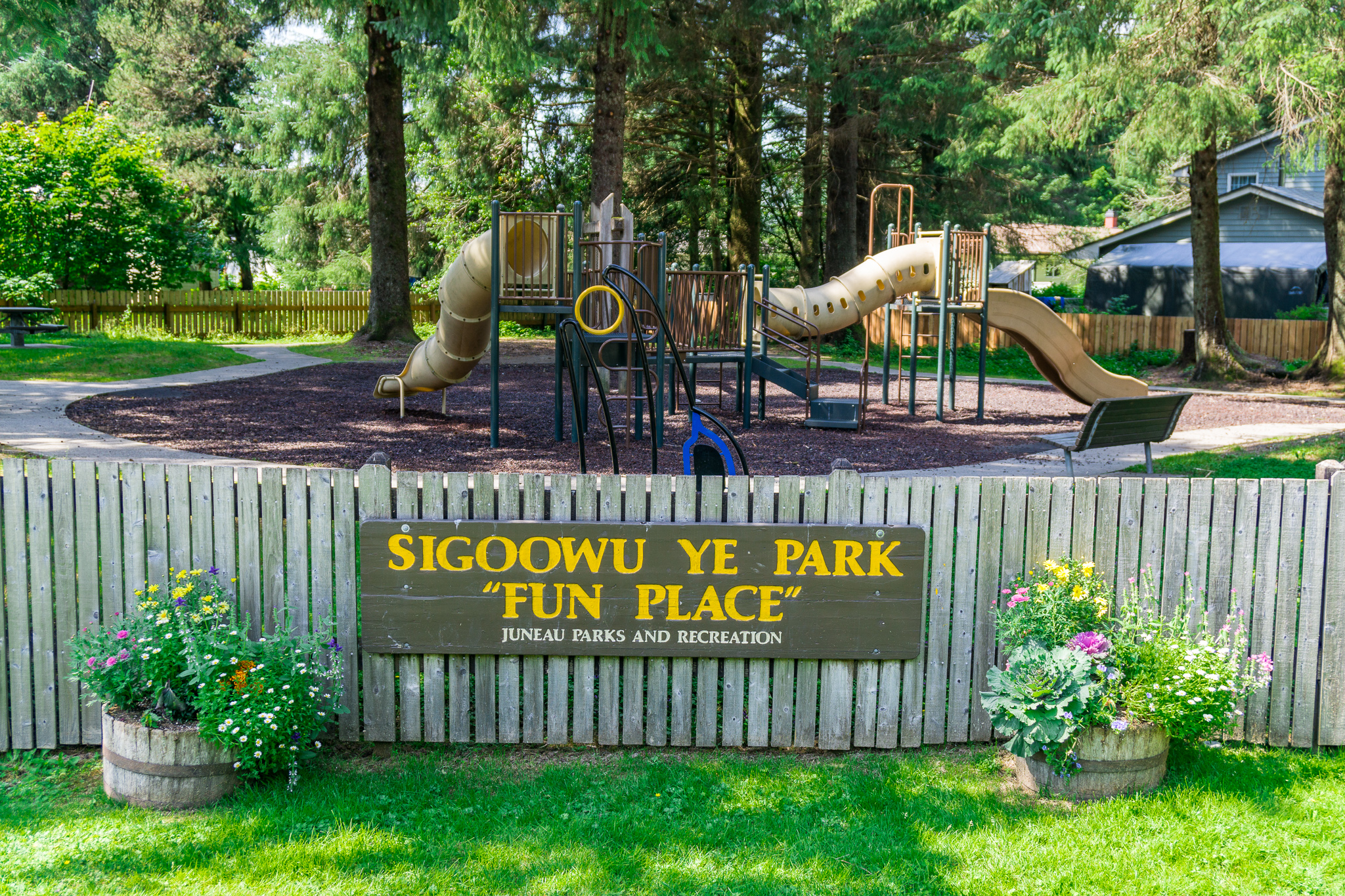 A playground surrounded by tall evergreen trees at Sigoowu Ye Park, also known as "Fun Place," in Juneau, Alaska. The fenced-in area features climbing structures, slides, and a soft mulch surface. A wooden sign on the fence reads “Sigoowu Ye Park ‘Fun Place’ – Juneau Parks and Recreation,” with colorful flower barrels on each side.