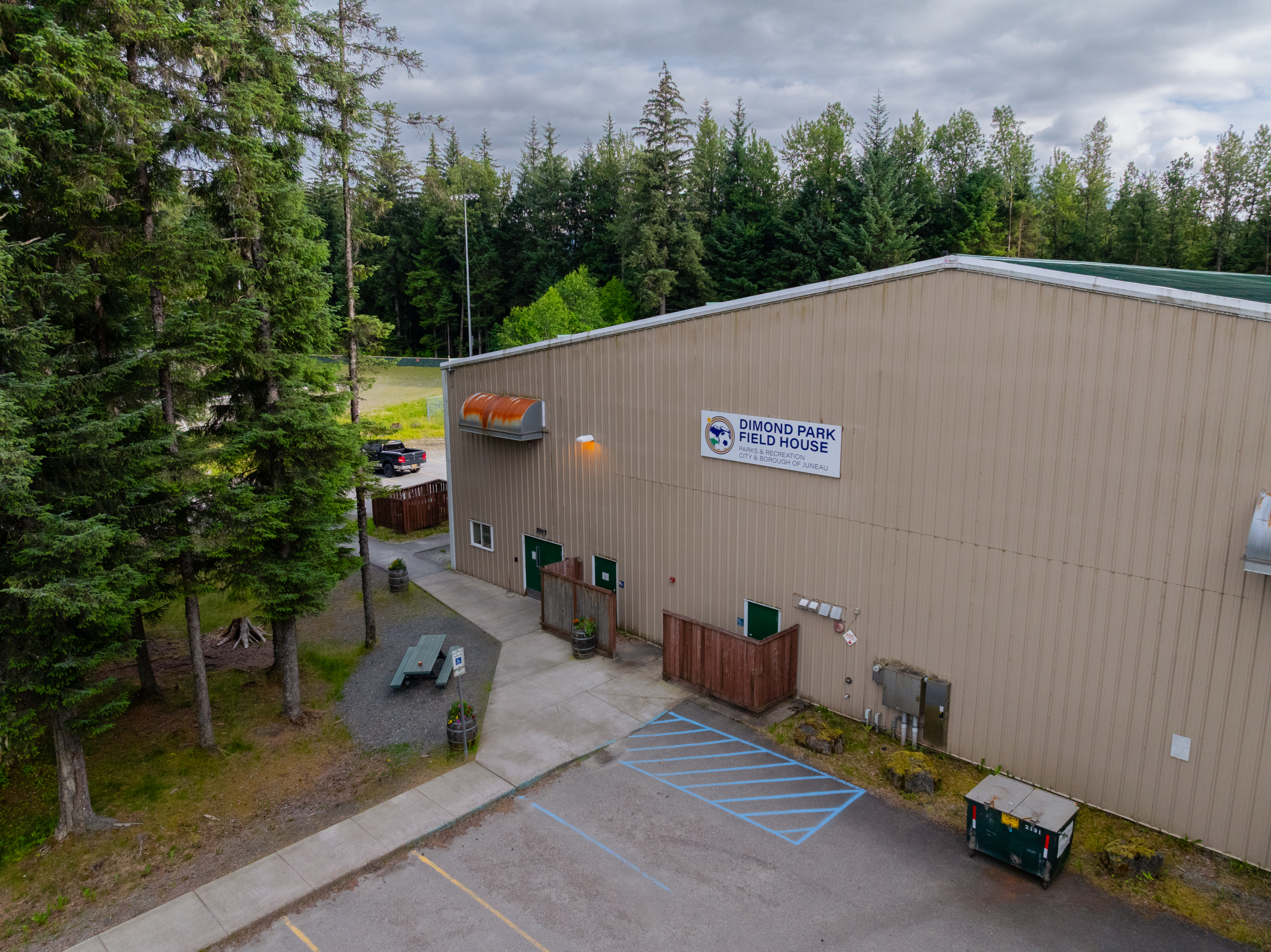 Exterior view of the Dimond Park Field House in Juneau, Alaska. Several cars are parked in the lot, and the building is surrounded by tall evergreen trees under an overcast sky. A sign reading 'Dimond Park Field House' is mounted on the tan metal building.