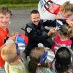 This is a picture of a Juneau Police Department officer crouching down among a group of children, smiling and helping to put a cardboard hat on a kid.