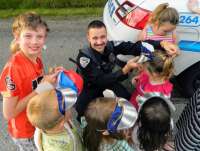 This is a picture of a Juneau Police Department officer crouching down among a group of children, smiling and helping to put a cardboard hat on a kid.