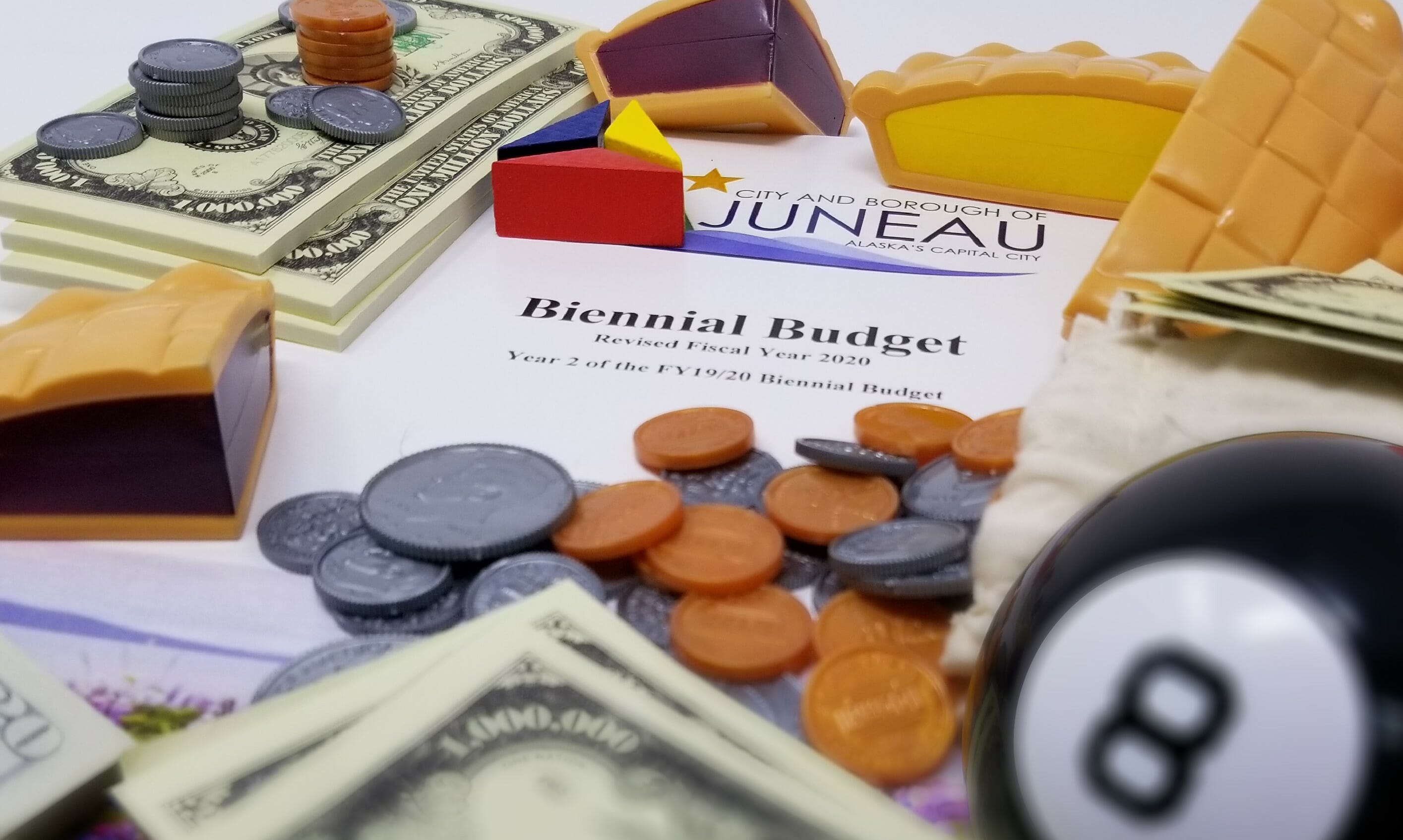 This is a staged image of the 2019 Budget Book amidst finance props like a Magic 8 ball, fake money, fake pie slices
