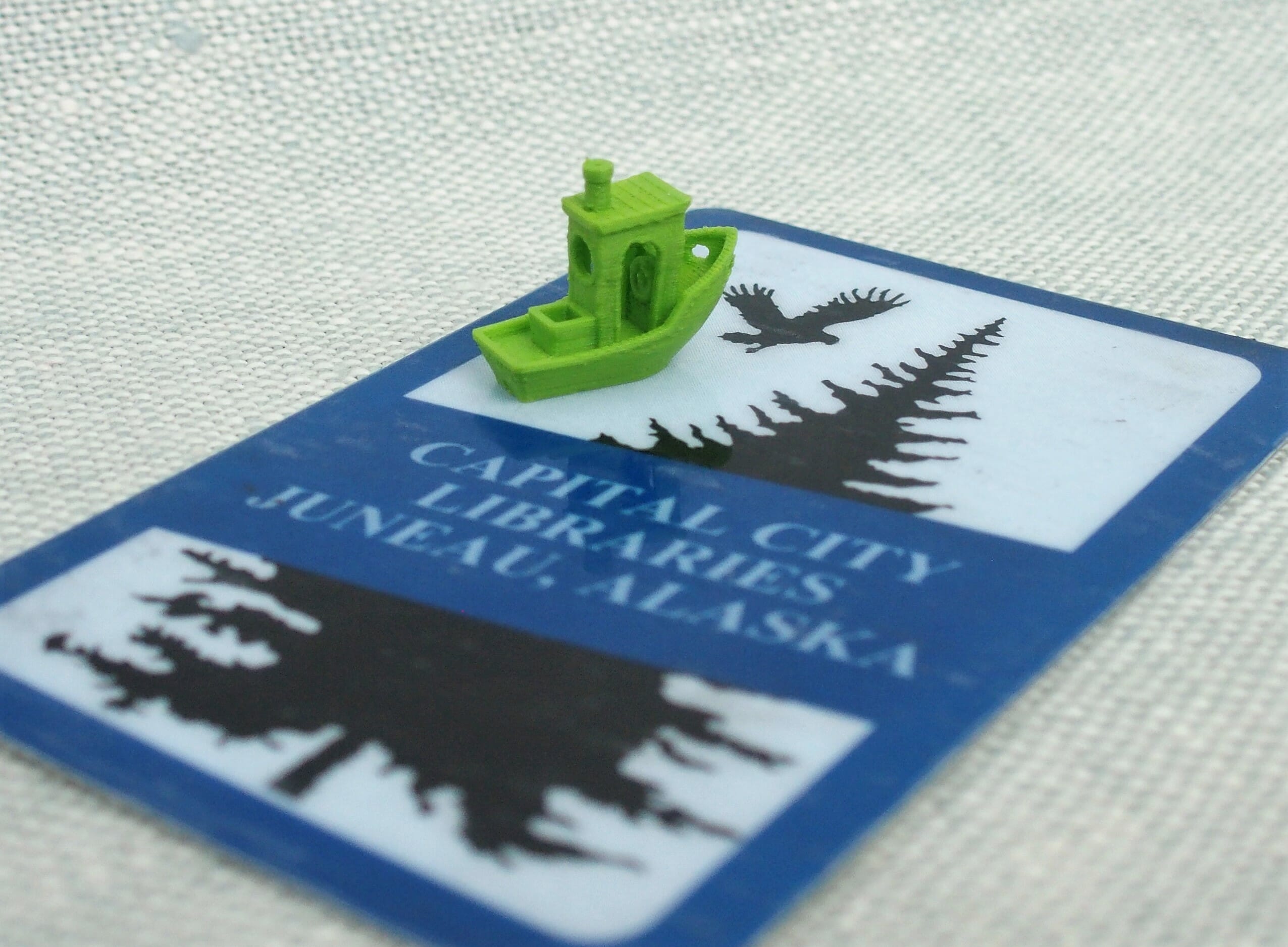 Photo of a small boat model prined in green PLA, sitting on top of a JPL library card. Visible details include a steering wheel inside the cabin and a hole in the center of the chimney. Layer lines and some blobbiness is visible on the walls of the cabin and under the gunnels.