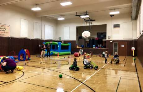 IMG_1909 Children riding bikes and playing with toys during Preschool Open Gym. Two dad stand in the background near the bouncy house.