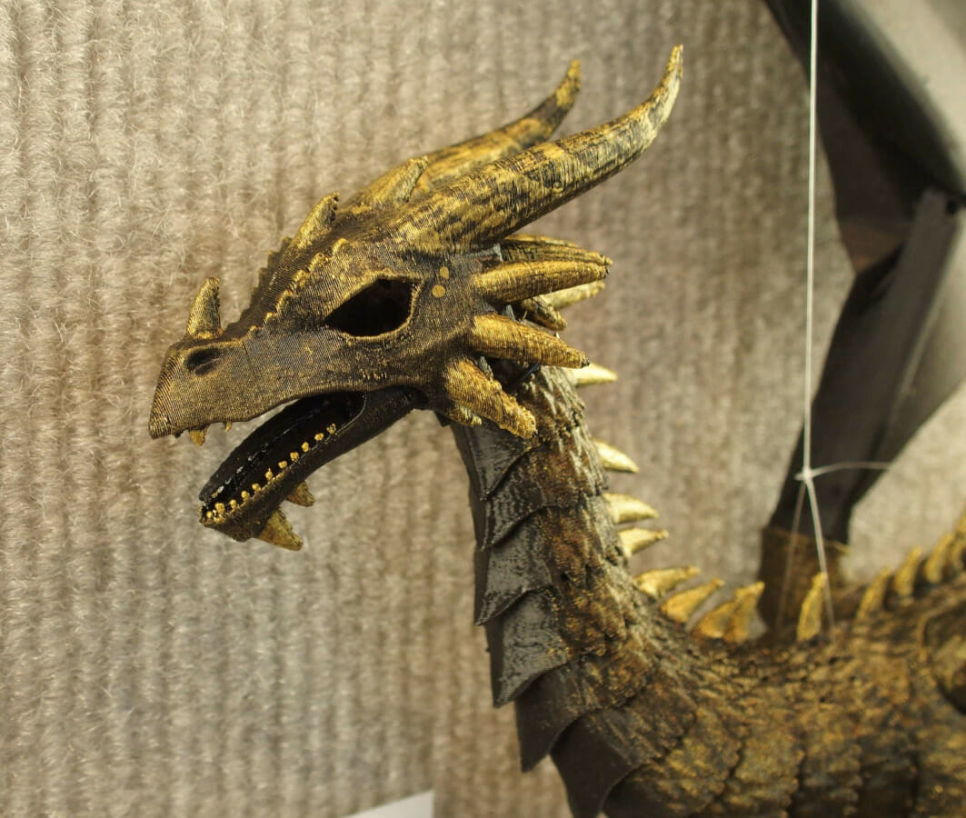 Head & shoulders photo of a 3D printed dragon. It is black plastic, brushed with gold on its face, back, horns, and teeth..