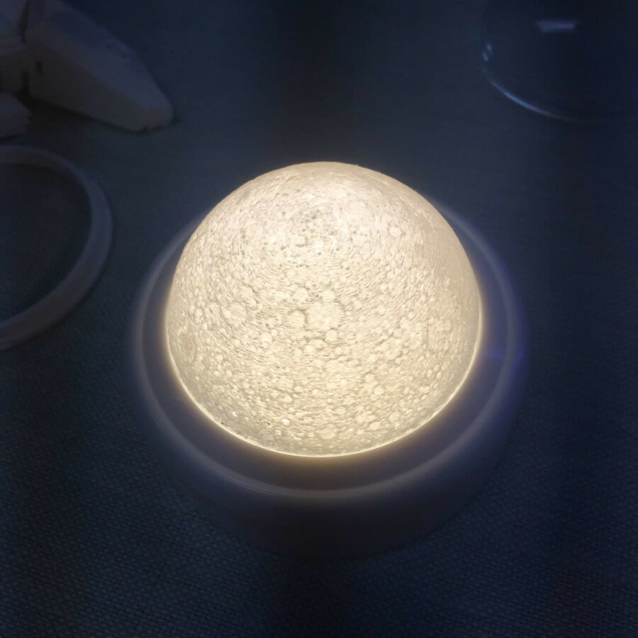 A white tap light about 3 1/2" in diameter, turned on. The original lens has been replaced by a 3D printed hemisphere textured like the surface of the moon. The plastic has variable thickness so the areas that are darker on the moon's surface are darker on the lamp as well.