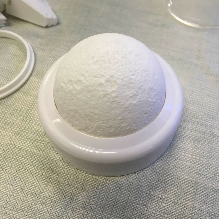 A white tap light about 3 1/2" in diameter. The original lens has been replaced by a 3D printed hemisphere textured like the surface of the moon.