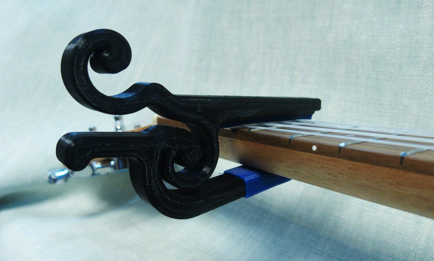Photo of a 3D printed capo, clamped onto the neck of a ukulele. The capo is black plastic, with a spiraled hinge and matching terminal on the fretboard-side jaw.