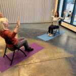 Photo of two women seated facing each other in chairs placed over yoga mats, each with both arms upraised
