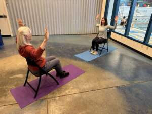Photo of two women seated facing each other in chairs placed over yoga mats, each with both arms upraised