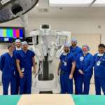 Photo of several smiling people in blue scrubs standing next to surgical robot