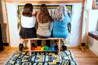 Back of view of three women seated side by side at a loom, with rainbow yarn displayed under the bench