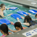Photo of swim instructor in the pool speaking to several young swimmers with kickboards