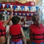 Photo of four children in red life vests standing poolside and listening to an instructor