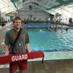 Photo of lifeguard standing in front of pool and holding red lifesaving device