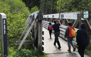 A group of four children, wearing backpacks and boots, cross a wooden bridge on the Fish Creek Trail. The bridge is made of weathered wood and metal supports, surrounded by lush green foliage. A sign on the bridge reads, "Pack it in, pack it out. Please pack out all trash." The children appear to be on a nature hike, with one holding a large stick while walking ahead.