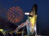 Nighttime photo of the whale statue lit up in several colors while in the background a large red firework explodes over Juneau.