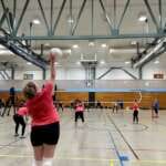 Photo from the back of a woman serving a volleyball toward players on the far side of the net