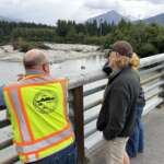 A small group, including a man in an Alaska DOT&PF safety vest, stand on a bridge and look over the Mendenhall River.