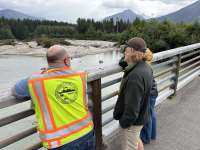 A small group, including a man in an Alaska DOT&PF safety vest, stand on a bridge and look over the Mendenhall River.