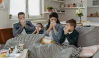 An unhappy-looking family sits wrapped in blankets on a couch, all blowing their noses