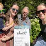 A multigenerational group of walkers smile beside a sign for Parks & Recreation Clue