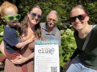 A multigenerational group of walkers smile beside a sign for Parks & Recreation Clue