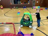 One small child pushes a dino-themed push car, with another child in the driver's seat.