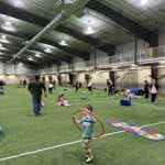 Young children play with hula hoops and other toys on the Field House turf as adults look on