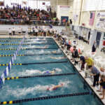 Competing swimmers approach the end of the pool as spectators crowding the bleachers cheer them on.