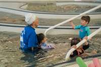 An aquatics instructor in swim cap guides three small children in a shallow pool.