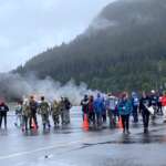 Dozens of volunteers gather with smoke and aircraft fire in the background