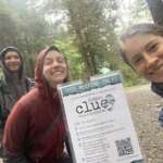 Three raingear-clad hikers pose for a selfie by a Parks & Rec Clue sign on the trail
