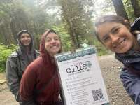 Three raingear-clad hikers pose for a selfie by a Parks & Rec Clue sign on the trail