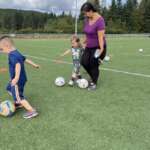 Two kids kick a soccer ball while with a guardian.