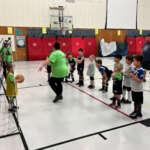 Two coaches and a group of children in an indoor gym next to a soccer net.