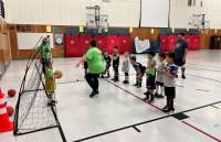 Two coaches and a group of children in an indoor gym next to a soccer net.