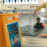 A swim instructor and two children in a pool next to a lane closed sign.