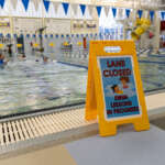 A sign that says "Lane Closed, Swim Lessons in Progress" sits on a pool deck in front of a lane. An instructor teaches kids how to float in the pool.