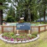 A park sign that says “Sigoowu Ye Park, Fun Place” stands above a bed of flowers. Behind the sign, a fenced park with new equipment is surrounded by trees.