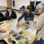 A group of four people sit around a table with cards while one person stands. The person standing holds a card in their hands, and one person sitting is arranging the cards on the table.
