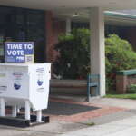 A ballot box stands next to a library building. On top of the ballot box is a blue sign that says "Time to Vote."