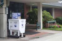 A ballot box stands next to a library building. On top of the ballot box is a blue sign that says 