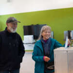 One person drops a ballot into a box as another person watches.