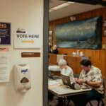 A doorway with a sign that says "Vote Here". Inside the door are election workers sitting at a table.