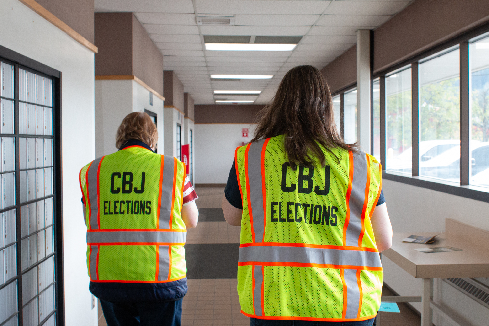 Two election workers in high visibility yellow vests walking away, down the USPS hallway with PO boxes to the left.